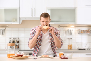 Photo of Young man eating tasty toasted bread at table in kitchen