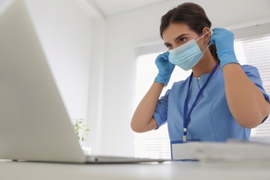 Photo of Doctor in medical gloves putting on protective mask at table in office