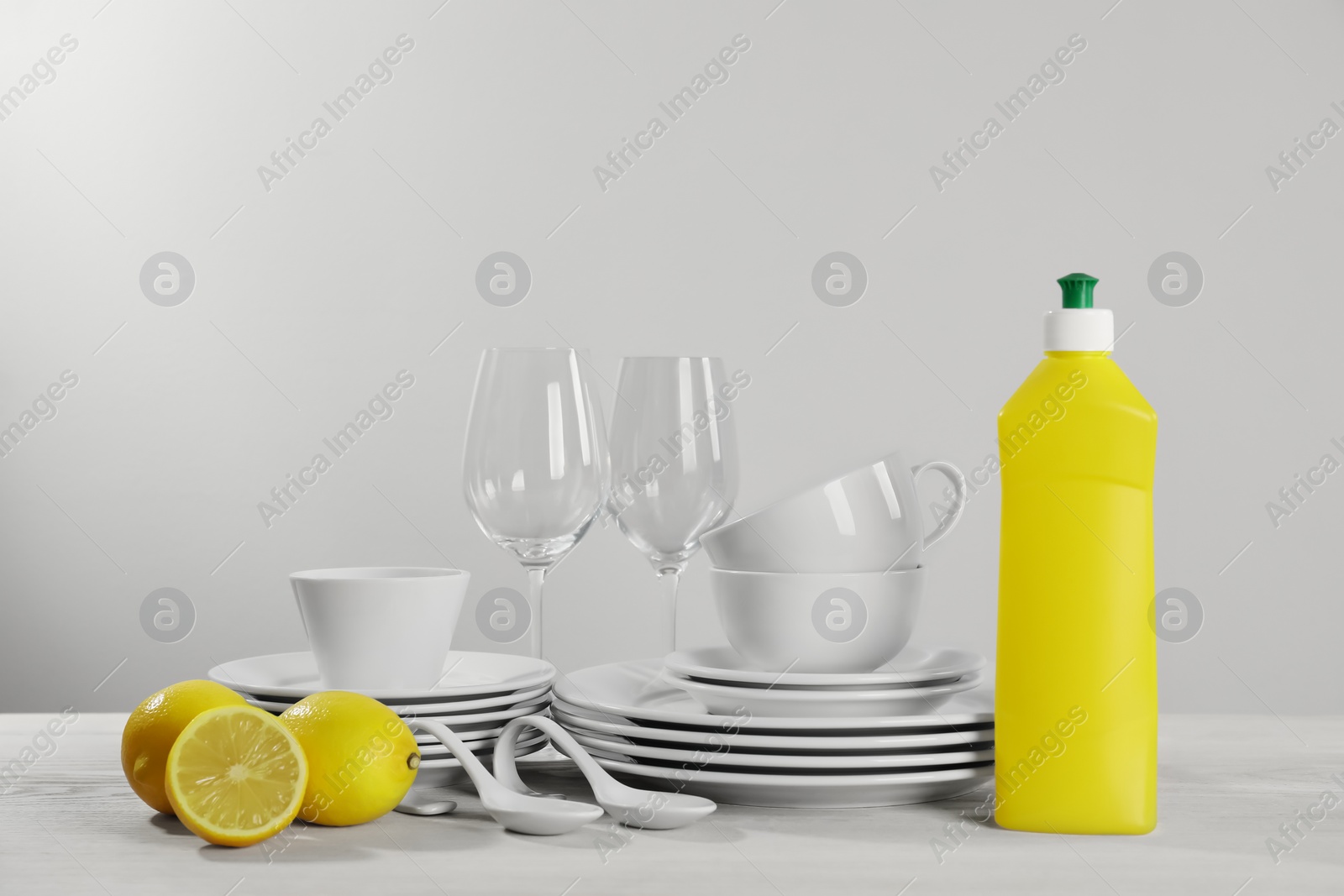 Photo of Set of clean tableware, glasses, dish detergent and lemons on white wooden table against light background