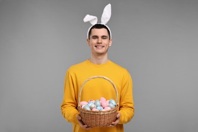 Photo of Easter celebration. Handsome young man with bunny ears holding basket of painted eggs on grey background