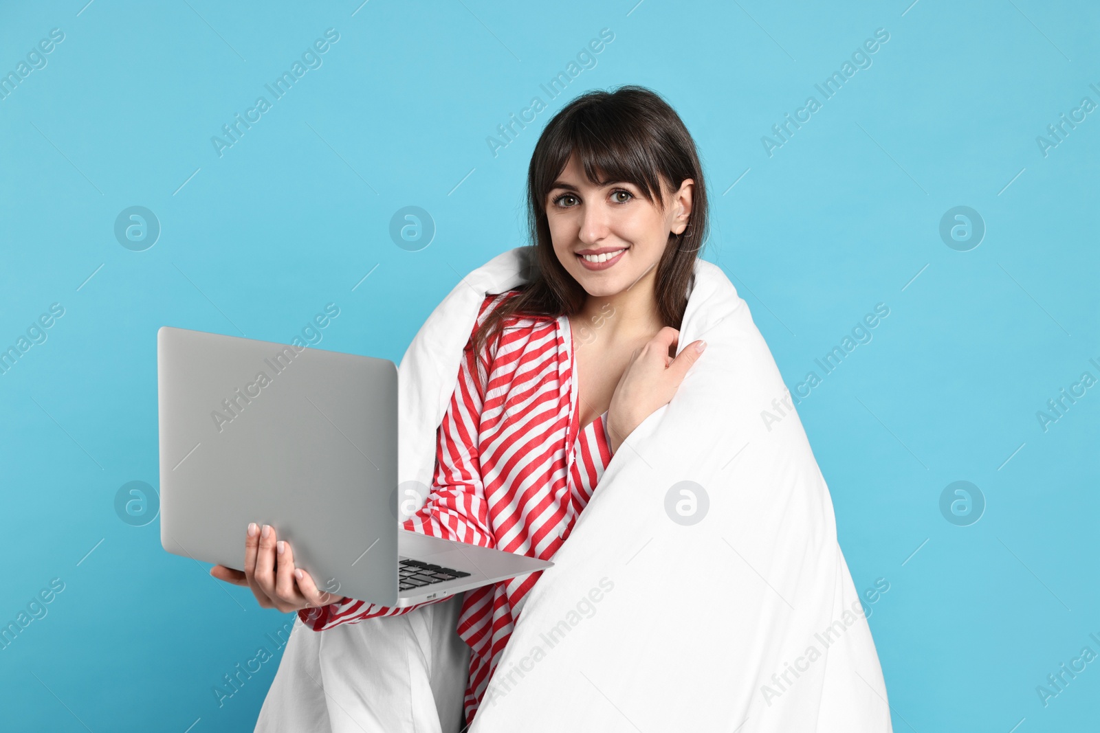 Photo of Happy woman in pyjama wrapped in blanket holding laptop on light blue background