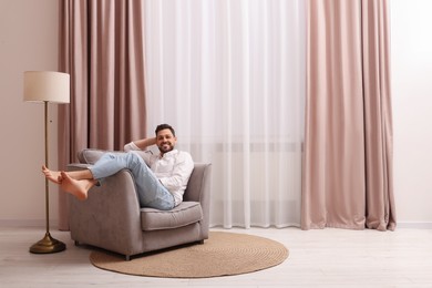 Happy man resting on armchair near window with beautiful curtains at home. Space for text