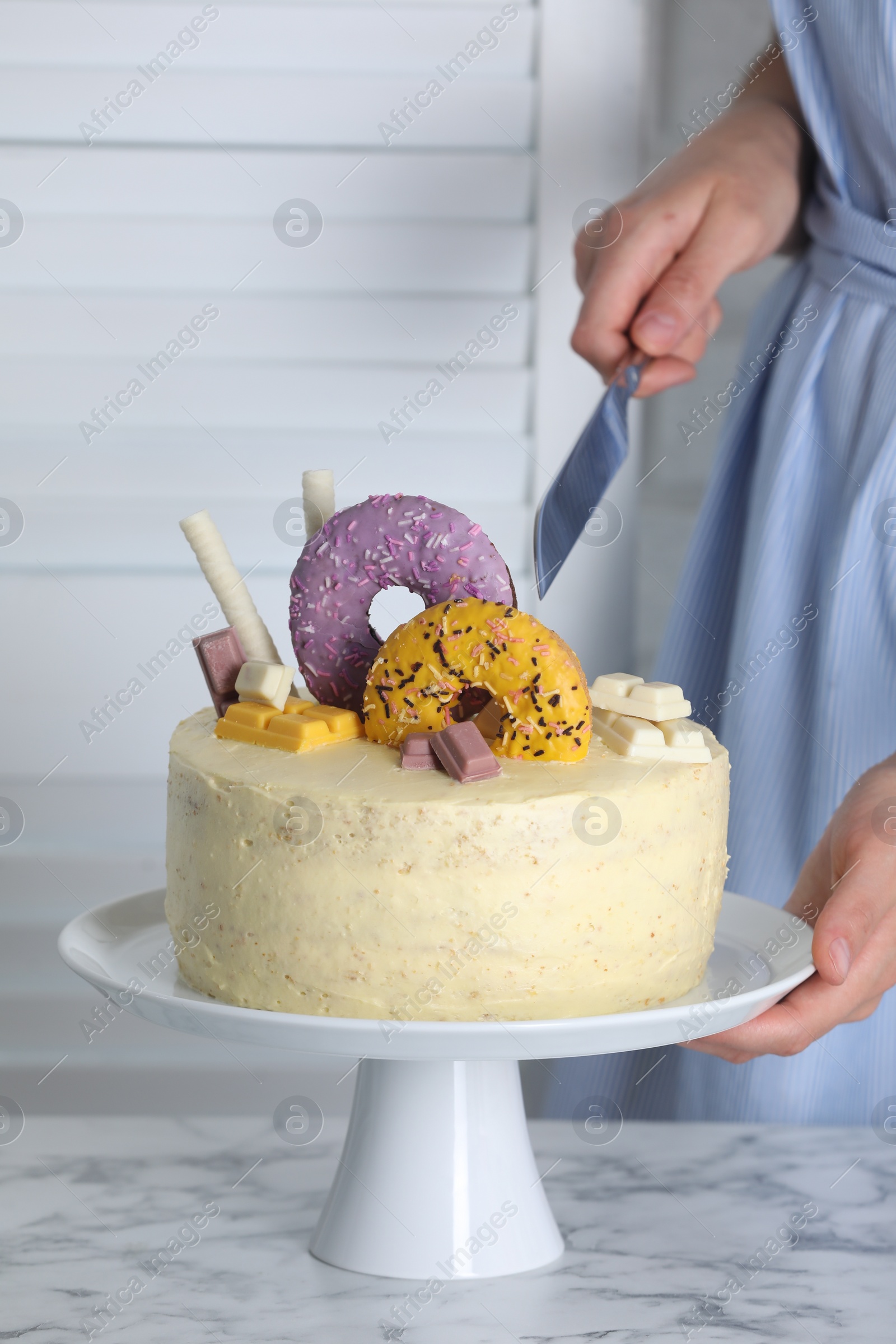 Photo of Woman cutting delicious cake decorated with sweets at white marble table, closeup