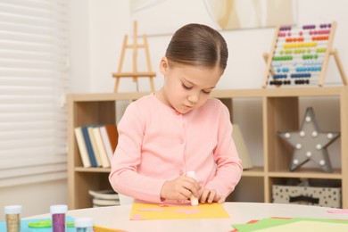 Photo of Cute little girl using glue stick at desk in room. Home workplace