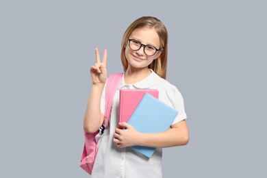 Cute girl in glasses with backpack and books on light grey background
