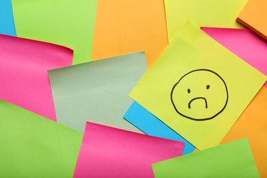 Photo of Sticky note with sad face among colorful paper, top view. Space for text