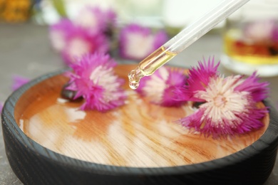 Photo of Dripping essential oil into bowl with water and flowers on table