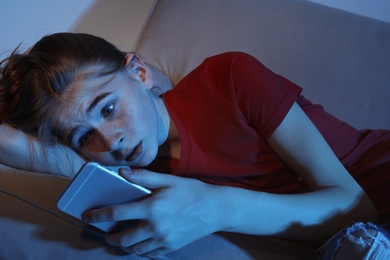 Photo of Shocked teenage girl with smartphone on sofa in dark room. Danger of internet