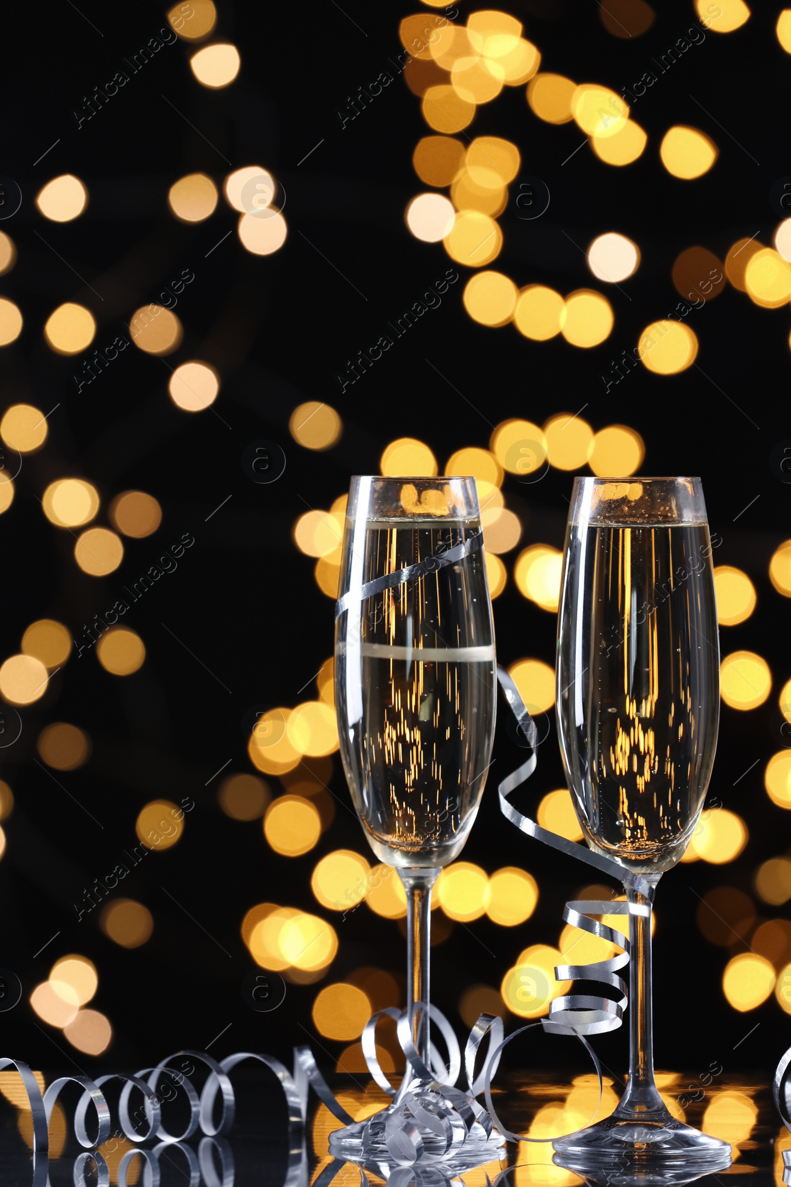 Photo of Glasses of champagne and serpentine streamers against black background with blurred lights. Space for text