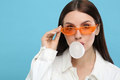 Beautiful woman in sunglasses blowing bubble gum on light blue background, space for text