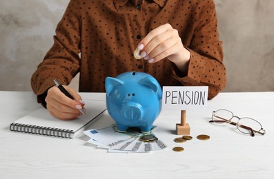 Photo of Pension savings. Woman writing in notebook and putting coin into piggy bank at white wooden table, closeup