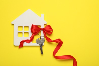 Photo of Wooden house model, red bow and key on yellow background, flat lay with space for text. Housewarming party