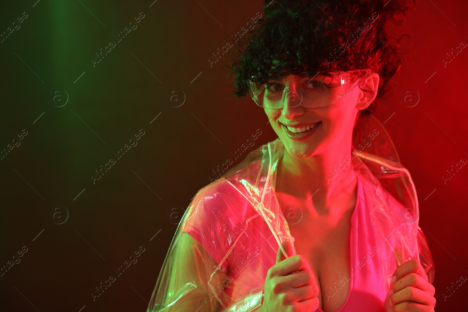 Photo of Beautiful young woman in transparent coat and sunglasses posing on color background in neon lights. Space for text