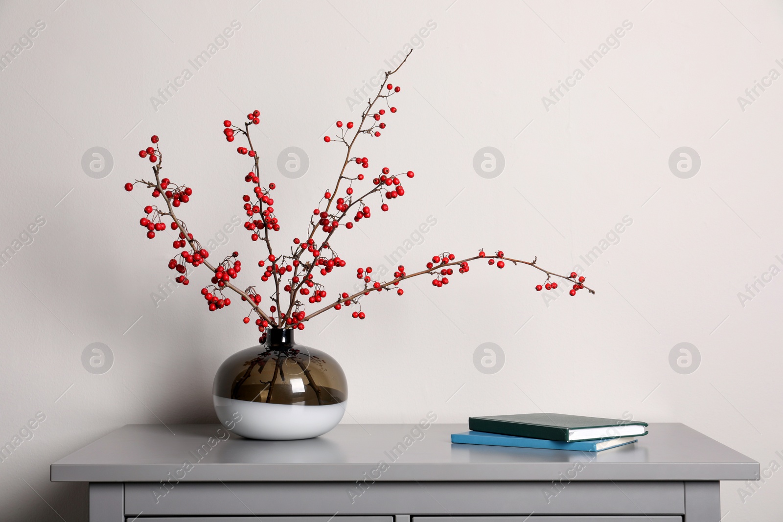 Photo of Hawthorn branches with red berries in vase and books on grey table indoors