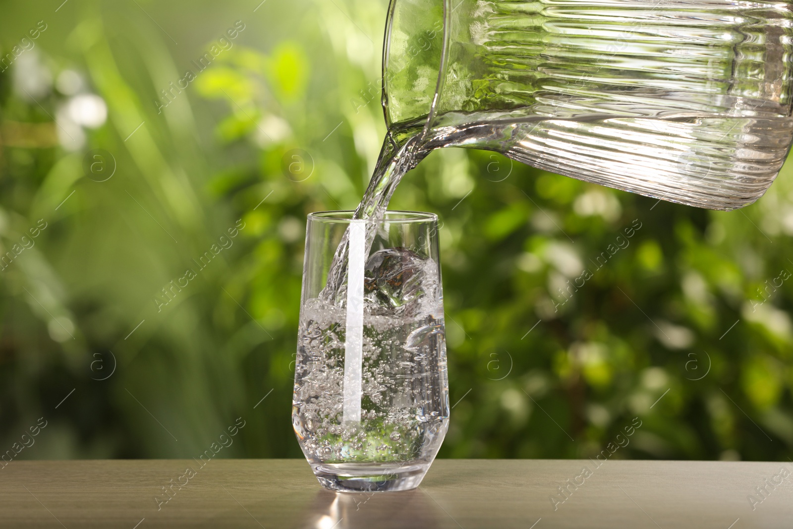 Photo of Pouring water from jug into glass on wooden table outdoors, closeup