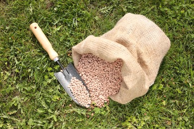 Photo of Granulated fertilizer in sack and shovel on green grass outdoors, top view