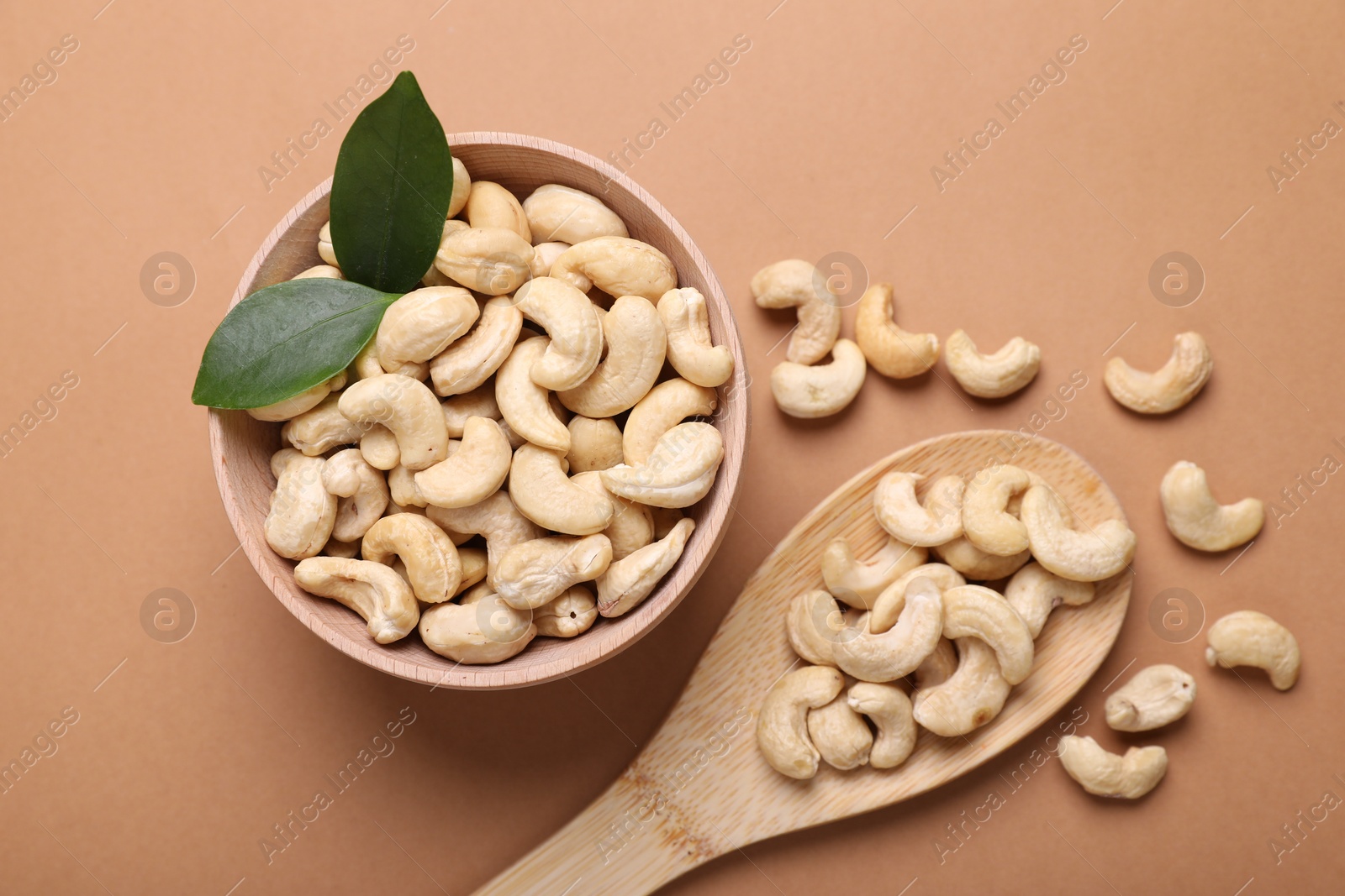 Photo of Tasty cashew nuts and green leaves on pale brown background, flat lay