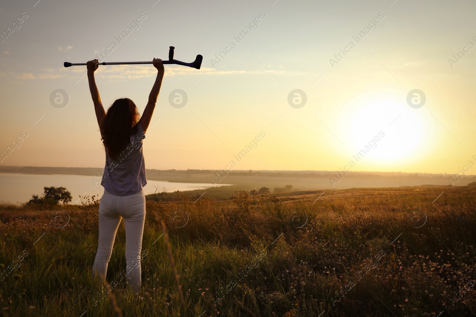 Photo of Woman raising hands with elbow crutch up to sky outdoors at sunrise, back view. Healing miracle