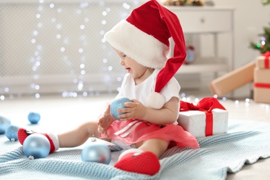 Photo of Cute baby in festive costume playing with Christmas decor on floor at home