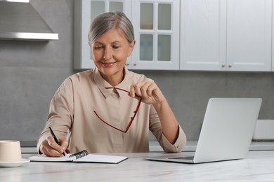 Photo of Beautiful senior woman taking notes near laptop at white marble table in kitchen