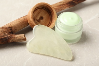 Photo of Jade gua sha tool, jar of cream and tree branch on grey table