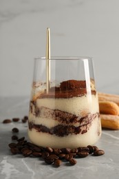 Photo of Tasty tiramisu in glass, coffee beans, spoon and biscuits on light grey table, closeup