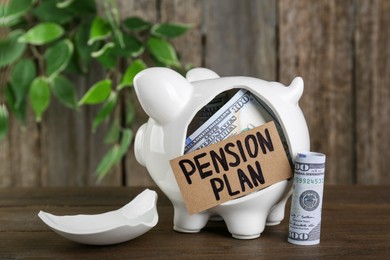 Photo of Card with phrase Pension Plan, broken piggy bank and dollar banknotes on wooden table. Retirement concept