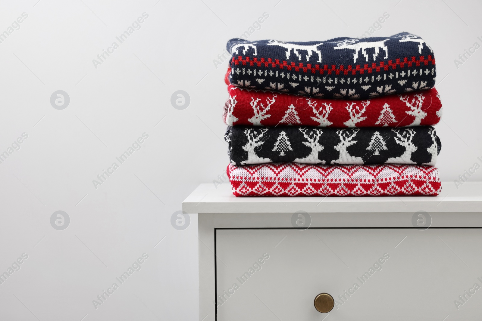 Photo of Different folded Christmas sweaters on chest of drawers against light background. Space for text