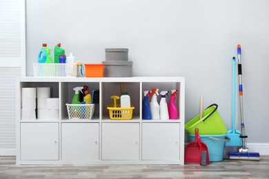 Photo of Shelving unit with detergents and cleaning tools near light grey wall indoors