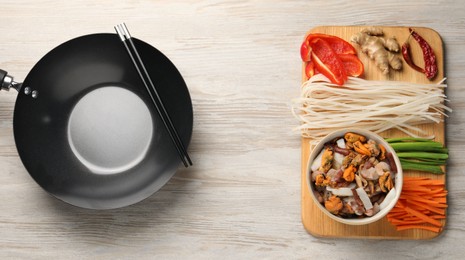 Black wok, chopsticks and board with products on light wooden table, top view