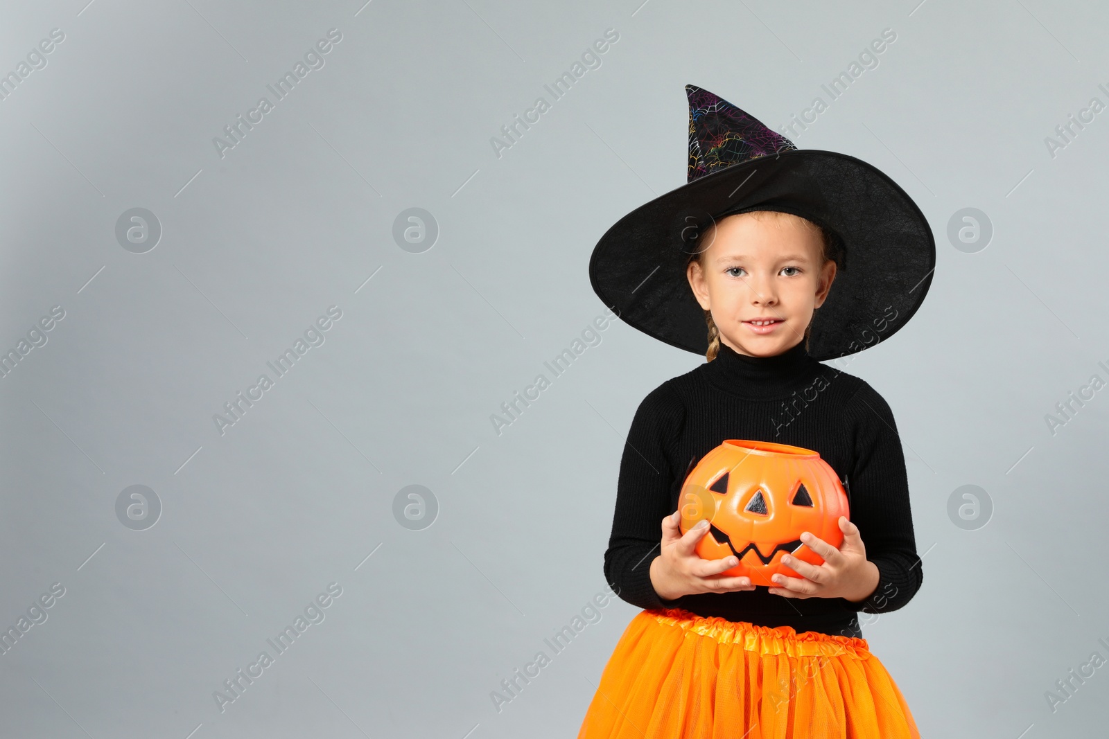 Photo of Cute little girl with pumpkin candy bucket wearing Halloween costume on grey background, space for text