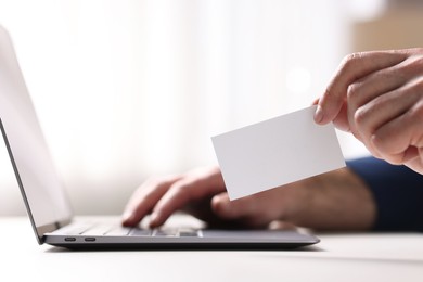 Photo of Man with laptop holding blank business card at white table indoors, closeup