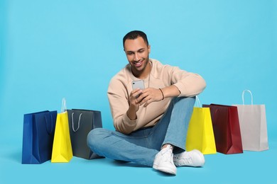 Happy African American man with shopping bags and smartphone on light blue background