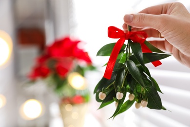 Photo of Woman holding mistletoe bunch with red bow indoors, closeup. Traditional Christmas decor