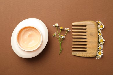 Photo of Jar of hair care cosmetic product, wooden comb and beautiful chamomile flowers on brown background, flat lay