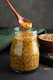 Photo of Taking whole grain mustard with spoon from jar on black wooden table