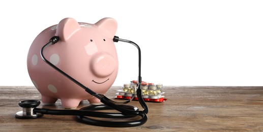 Piggy bank with stethoscope and pills on wooden table against white background, space for text. Medical insurance