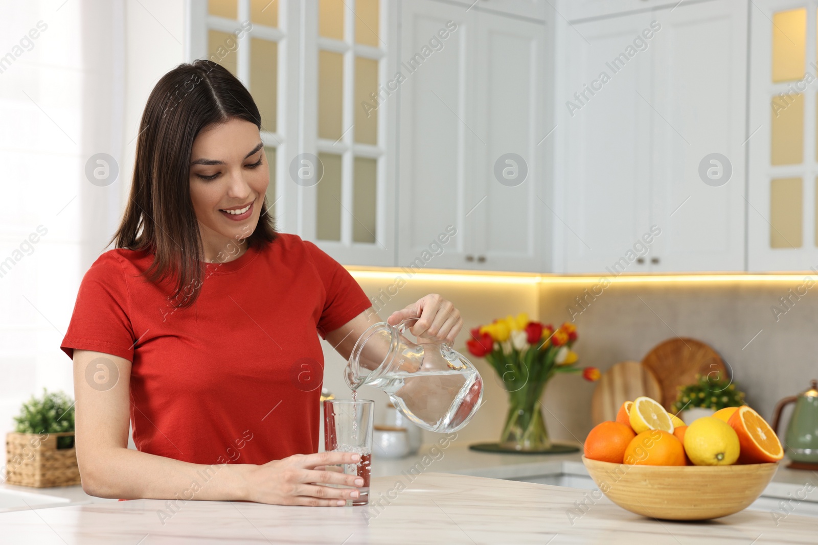 Photo of Young woman pouring water into glass from jug in kitchen