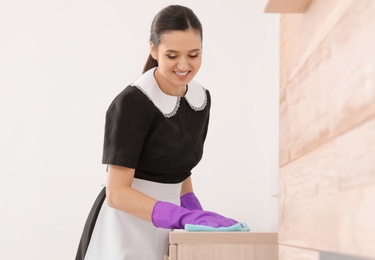 Photo of Young maid dusting furniture with rag in hotel room