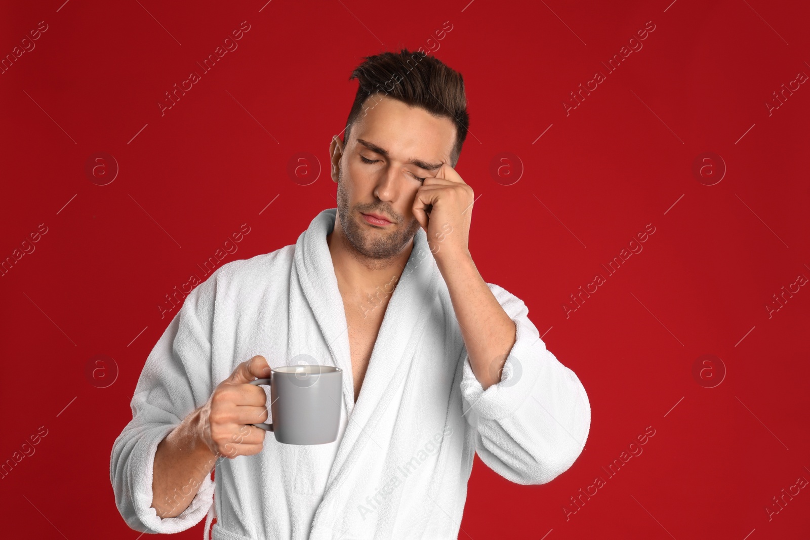 Photo of Sleepy young man in bathrobe  with cup of coffee on red background