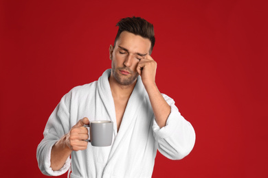 Sleepy young man in bathrobe  with cup of coffee on red background