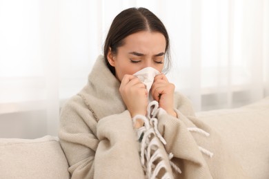 Sick woman wrapped in blanket with tissue blowing nose on sofa at home. Cold symptoms