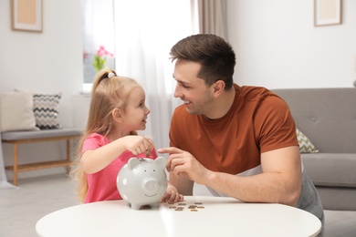 Photo of Father and daughter putting coin into piggy bank at table indoors. Saving money