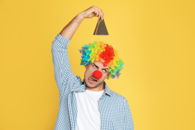 Photo of Funny man with clown nose, party hat and rainbow wig on yellow background. April fool's day