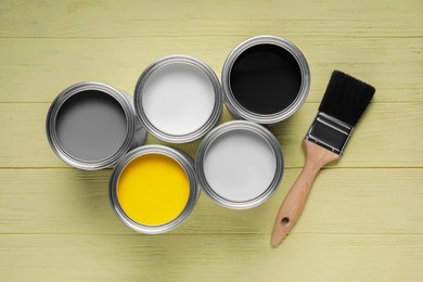 Cans of white, black, yellow and grey paints with brush on olive wooden table, flat lay
