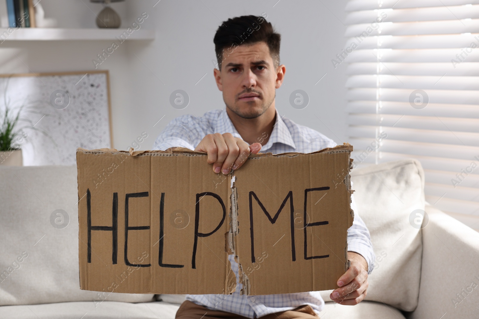 Photo of Unhappy man with HELP ME sign on sofa indoors