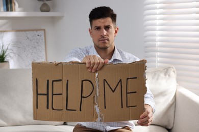 Photo of Unhappy man with HELP ME sign on sofa indoors