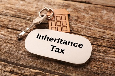 Photo of Inheritance Tax. Card and key with key chain in shape of house on wooden table, closeup