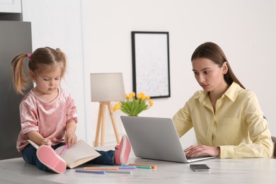 Photo of Mother with laptop working remotely while her daughter drawing at home. Child sitting on desk