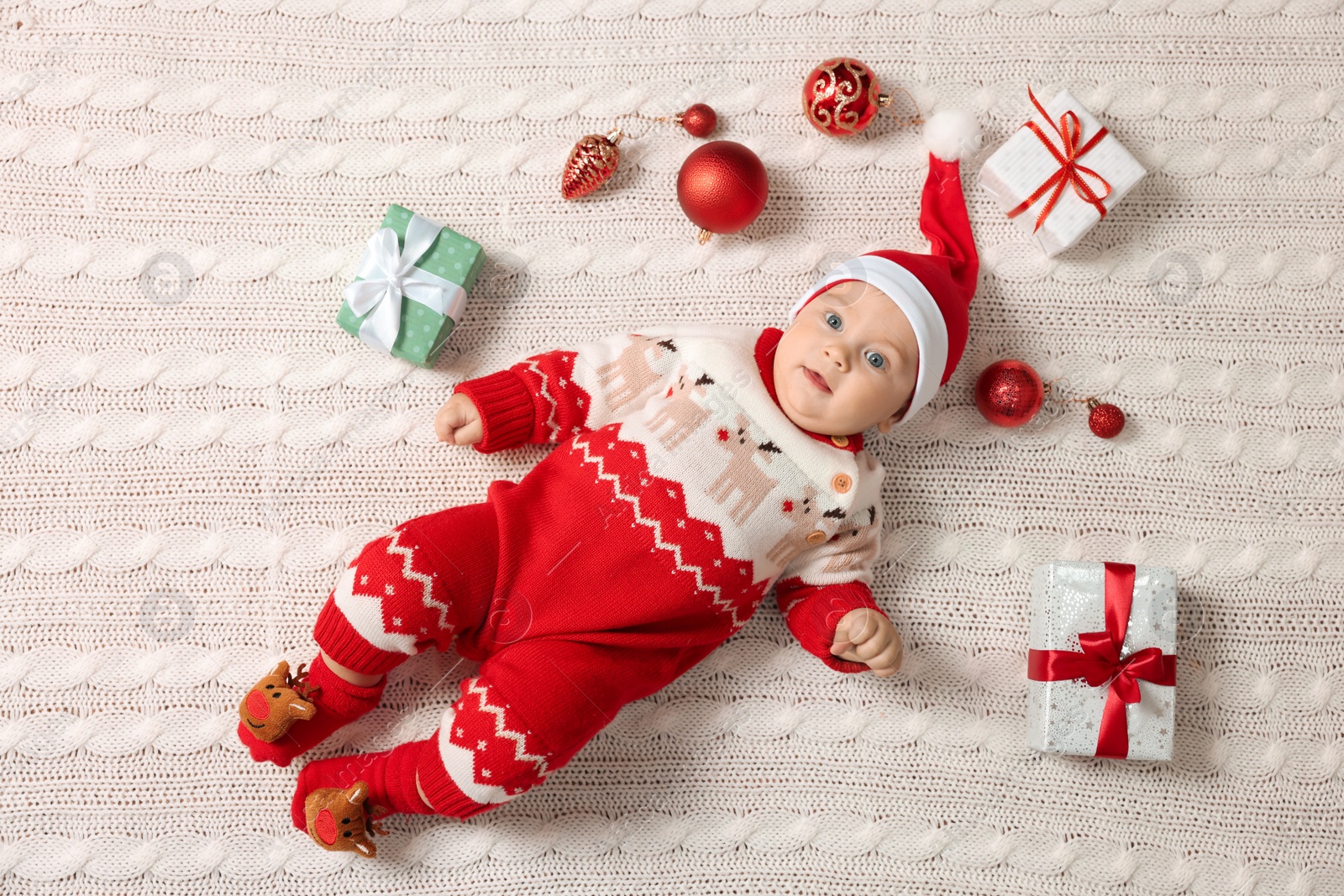 Photo of Cute little baby in Christmas outfit surrounded by festive items on white knitted plaid, top view. Winter holiday